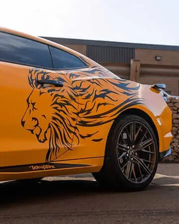 Wraptors Orlando, Vehicle Wraps, PPF & Tint A yellow car featuring a large black lion graphic on the side, wrapped with precision by Wraptors Orlando, is parked outside a building with stone and metal accents. This striking design showcases the artistry of Orlando Florida Car Wraps. Orlando, FL