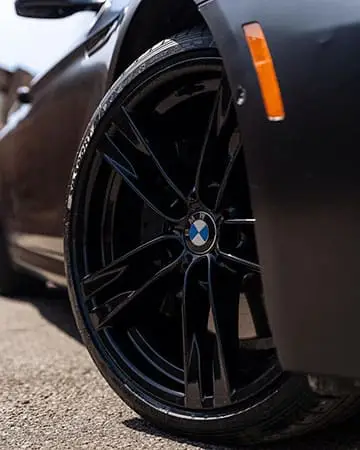 Wraptors Orlando, Vehicle Wraps, PPF & Tint Close-up of a black BMW car wheel and tire, showing the BMW logo on the wheel hub and part of the car’s black body and orange side reflector, enhanced with premium PPF protection by Wraptors Orlando—your trusted experts for Orlando Florida Car Wraps. Orlando, FL