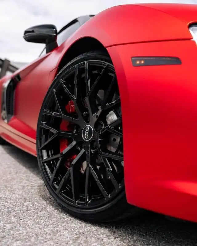 Wraptors Orlando, Vehicle Wraps, PPF & Tint Close-up view of a black alloy wheel on a matte red Audi sports car, featuring a premium finish by Wraptors Orlando. The vehicle, parked on a paved surface with the door slightly open, showcases the quality and precision of Orlando Florida Car Wraps and PPF application. Orlando, FL