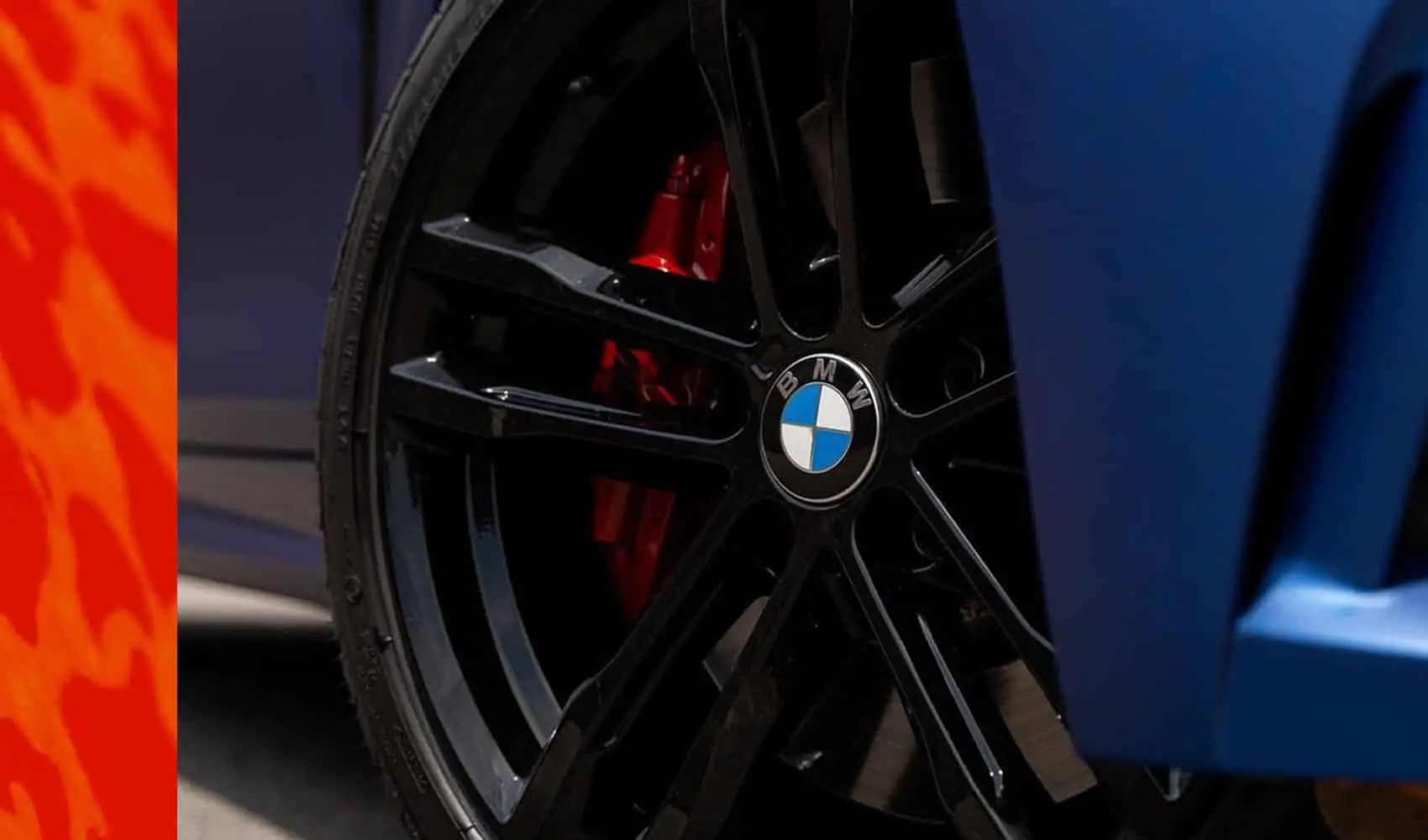 Wraptors Orlando, Vehicle Wraps, PPF & Tint Close-up of a black BMW wheel rim with the BMW logo in the center and a red brake caliper behind it, professionally detailed by Wraptors Orlando. Part of a blue car body, protected with premium PPF, and an orange surface are also visible—an example of the exceptional Orlando Florida Car Wraps expertise. Orlando, FL