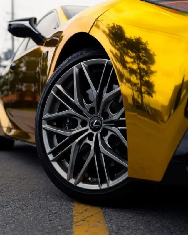 Wraptors Orlando, Vehicle Wraps, PPF & Tint Close-up of a Lexus sports car’s front wheel and tire, with a reflective gold exterior—expertly finished by Wraptors Orlando—showing trees and buildings in the paint. Featuring premium Orlando Florida Car Wraps and PPF protection, the car is parked by a yellow street line. Orlando, FL