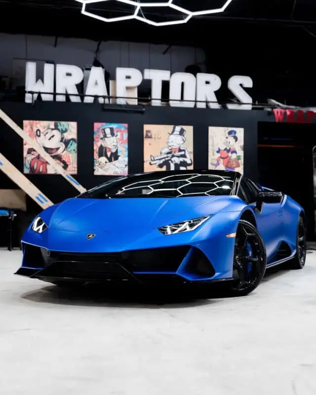 Wraptors Orlando, Vehicle Wraps, PPF & Tint A blue Lamborghini sports car, protected with premium PPF and expertly finished by Wraptors Orlando, is parked indoors with cartoon art—including Mickey Mouse and Scrooge McDuck—displayed on the wall behind it. This scene perfectly captures the vibrant style synonymous with Orlando Florida Car Wraps. Orlando, FL