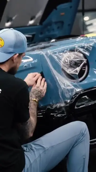 Wraptors Orlando, Vehicle Wraps, PPF & Tint A person wearing a blue cap applies a transparent protective film, also known as PPF, to the front of a blue car near the headlight—showcasing expert attention to detail from Wraptors Orlando, a leader in Orlando Florida Car Wraps. Orlando, FL