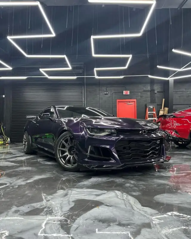 Wraptors Orlando, Vehicle Wraps, PPF & Tint A black sports car featuring a sleek PPF finish is parked indoors on a glossy floor under geometric ceiling lights, with a red door and ladders visible in the background—a testament to the quality and style offered by Wraptors Orlando, your experts for Orlando Florida Car Wraps. Orlando, FL